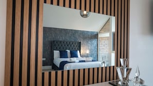 Una habitación de hotel moderna con un espejo grande, paneles de madera a rayas, un cabecero capitonado y una mesita de noche con una lámpara y cristalería.