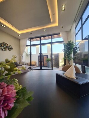 A modern living room with a large window, a sofa, and a potted plant. - Villa Bali Jeddah (Jeddah)