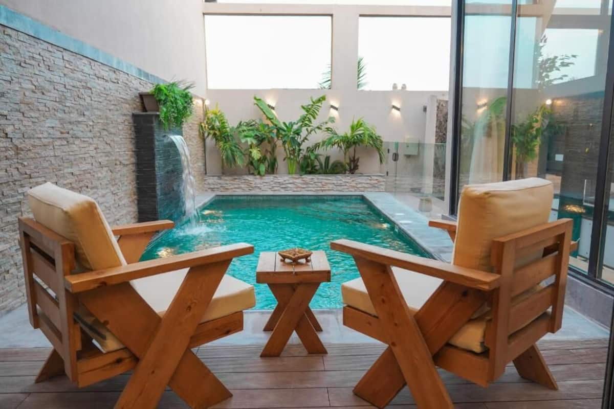 A pool area with wooden chairs and a table, surrounded by plants and a stone wall.
