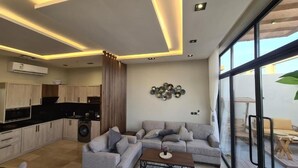 Una sala de estar moderna con un sofá gris, una mesa de centro y una zona de cocina con armarios de madera y un equipo de aire acondicionado.