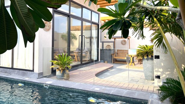 Un área de piscina moderna al aire libre con un porche acristalado, terraza de madera y exuberante vegetación tropical.