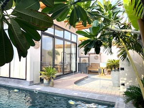 A modern outdoor pool area with a glass-enclosed patio, wooden deck, and lush tropical plants.