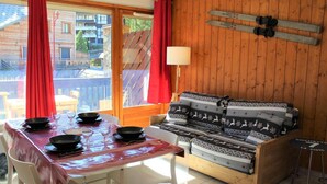 Um quarto aconchegante com um sofá de madeira, uma mesa de jantar posta com pratos e copos, e uma janela com vista para um edifício.