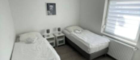 A small, modern bedroom with two single beds, a nightstand, a bedside table, and a window with blinds.