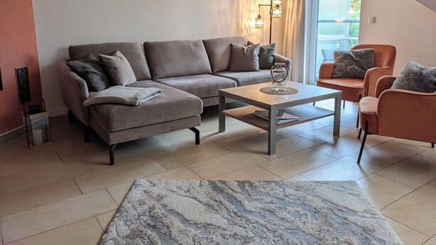 A modern living room with a sectional sofa, a coffee table, and a small side table. There is a large window with curtains, a wall-mounted light fixture, and a decorative wall piece. - Traumhafte Wohnung Über Zwei Ebenen mit Infrarotkabine, Balkon und Terrasse (Göhren-Lebbin)