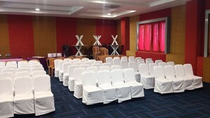 A conference room with rows of white chairs, a stage with a red curtain backdrop, and a carpeted floor. - Hotel Woodside Prestige Tirupati (Tirupati)