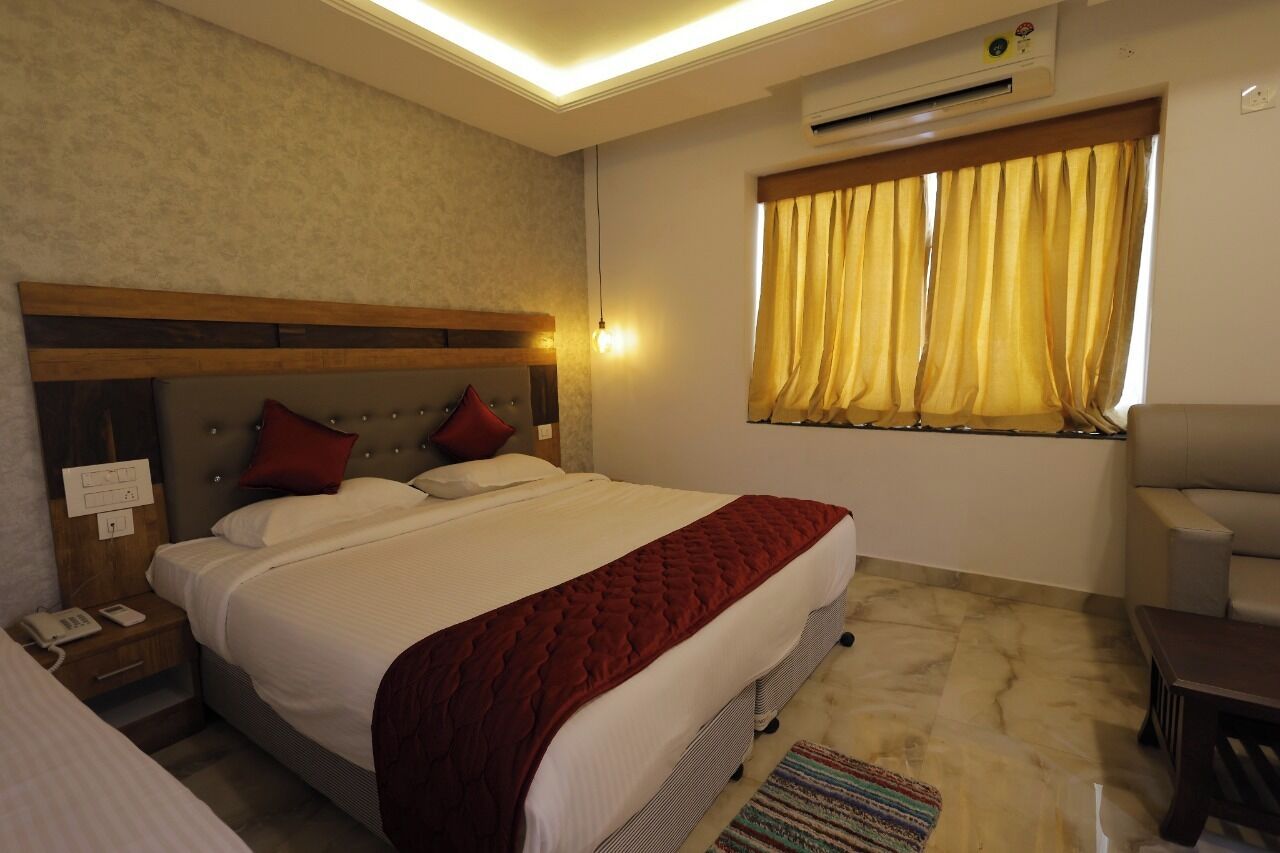 A hotel room with a bed, bedside table, lamp, air conditioning unit, window with curtains, and a chair.