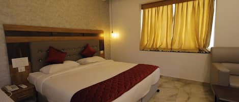 A hotel room with a bed, bedside table, lamp, air conditioning unit, window with curtains, and a chair.
