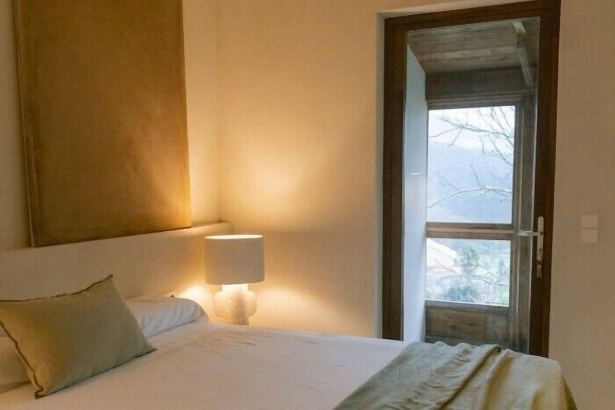 Un dormitorio con una cama, una mesita de noche con lámpara y una ventana con vista a árboles.