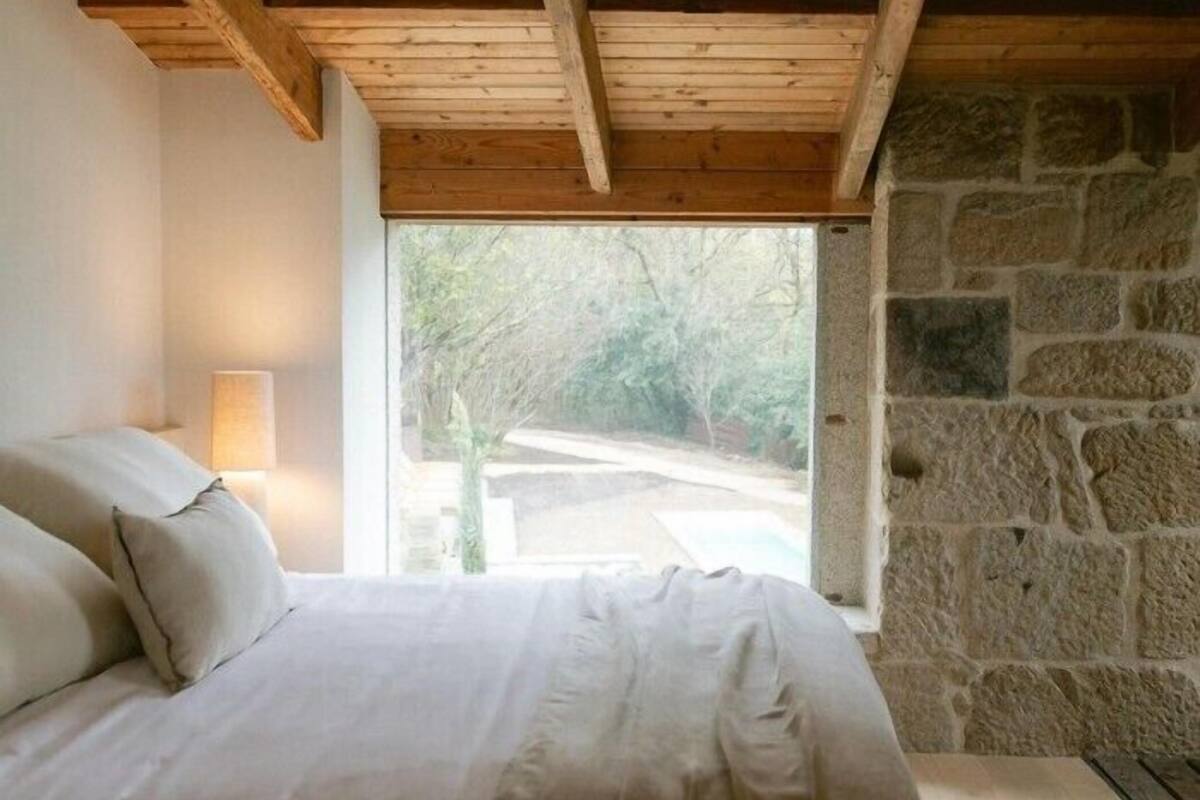 Un dormitorio con pared de piedra, cama con ropa de cama blanca y ventana con vistas al verdor.