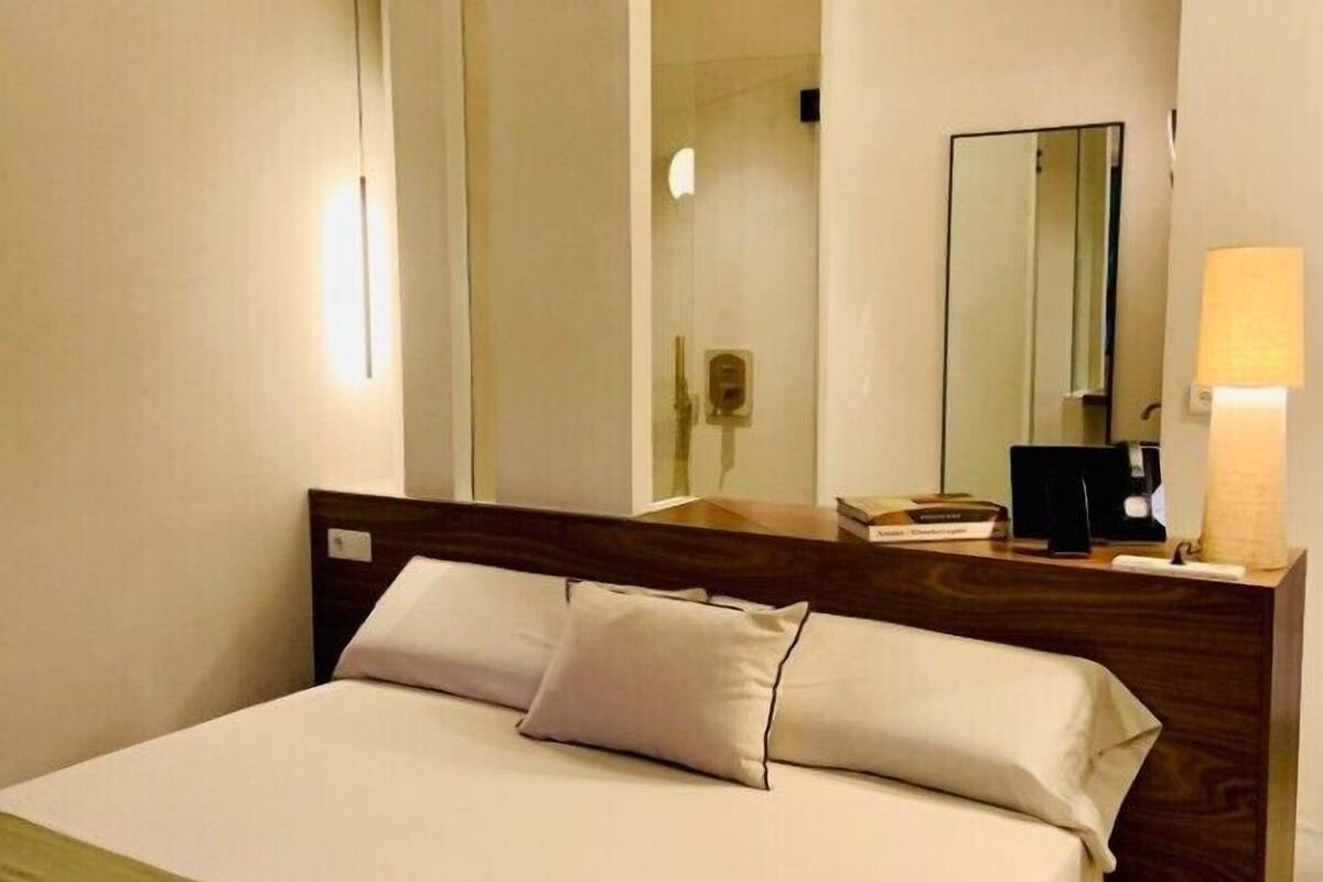 Habitación de hotel con cama, mesita de noche, espejo y lámpara de pared.