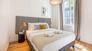 A neatly made bed with a checkered headboard, two bedside lamps, and a window with curtains. - Sweett | Wagram Cosy (Paris)