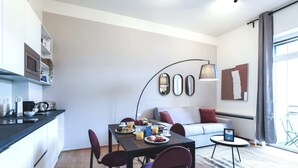 En moderne stue med spisebord, stole, sofa og sofabord. Der er et tekøkken med mikrobølgeovn og elkedel. Værelset har et stort vindue med gardiner og et spejl monteret på væggen.