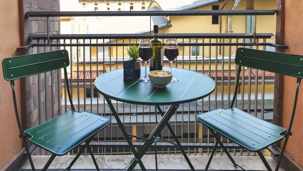 En lille balkon med et rundt bord, to grønne stole, en skål chips, to vinglas og en vinflaske.