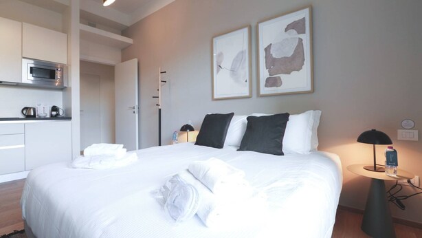 A hotel room with a bed, a bedside table with a lamp, a water bottle, and a microwave in the kitchenette. - Sweett | Classy Duomo II (Milano)