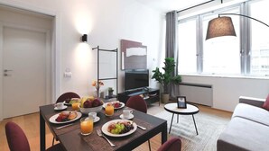Et moderne hotelværelse med spiseplads, sofa, tv og et stort vindue med gardiner.