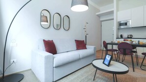 En moderne stue med en hvid sofa, røde puder, et rundt sofabord og en gulvlampe. Et køkken med hvide skabe og en mikrobølgeovn ses i baggrunden.