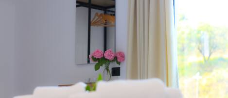 A hotel room with a bed, a vase with pink flowers, and a mirror reflecting a wardrobe.
