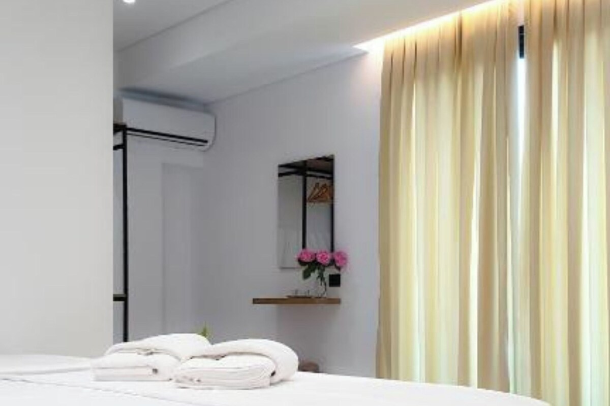 A modern bedroom with a large bed, a bedside table with a mirror, and a window with curtains.