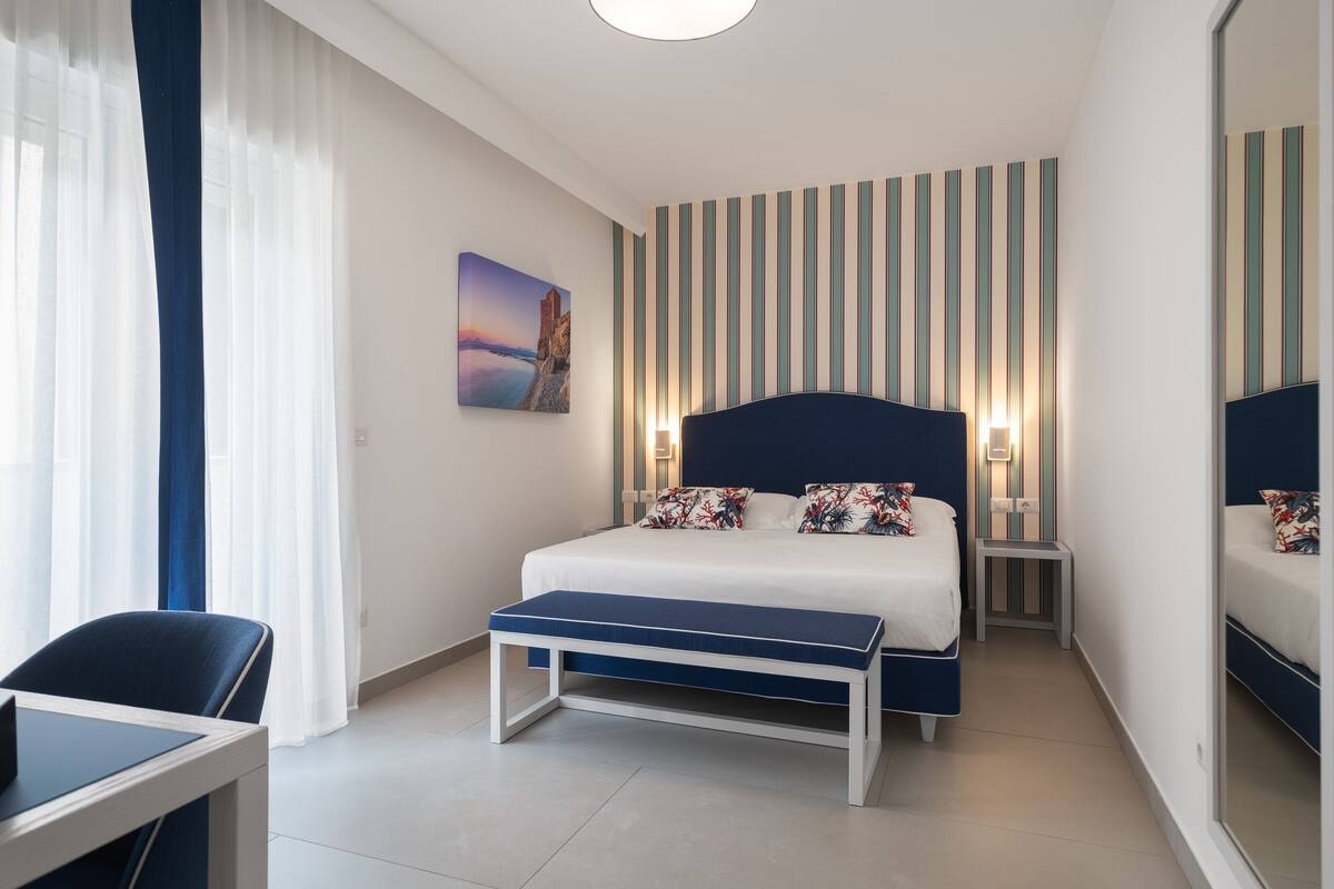 A modern hotel room with a large bed, bedside tables, a bench, a desk, and a large mirror.