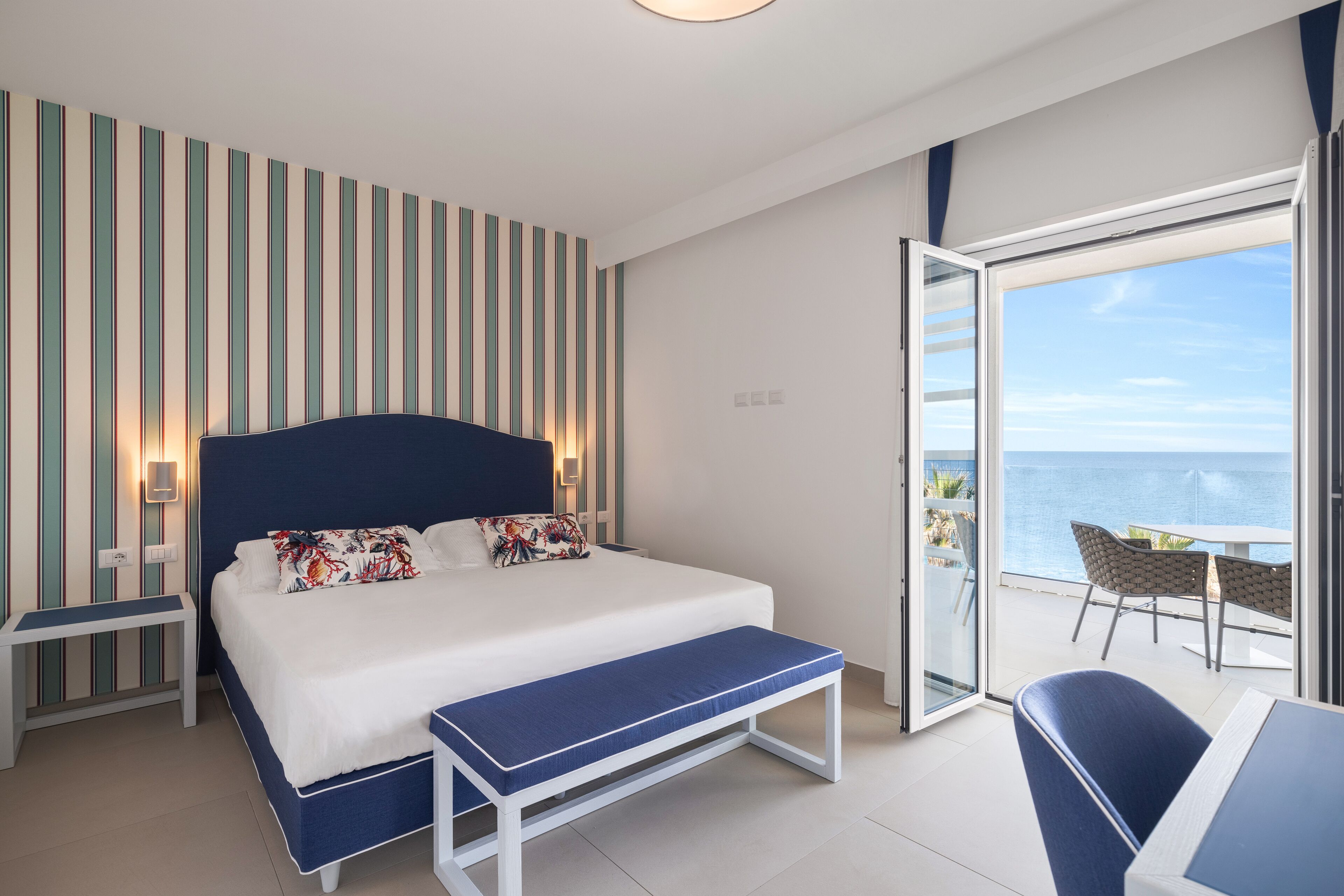 A bedroom with a bed, bench, bedside table, and a view of the ocean through a sliding door.