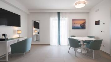 Uma sala de estar moderna com televisão de tela plana, uma mesa de jantar com cadeiras, máquina de café e uma janela grande com cortinas translúcidas.
