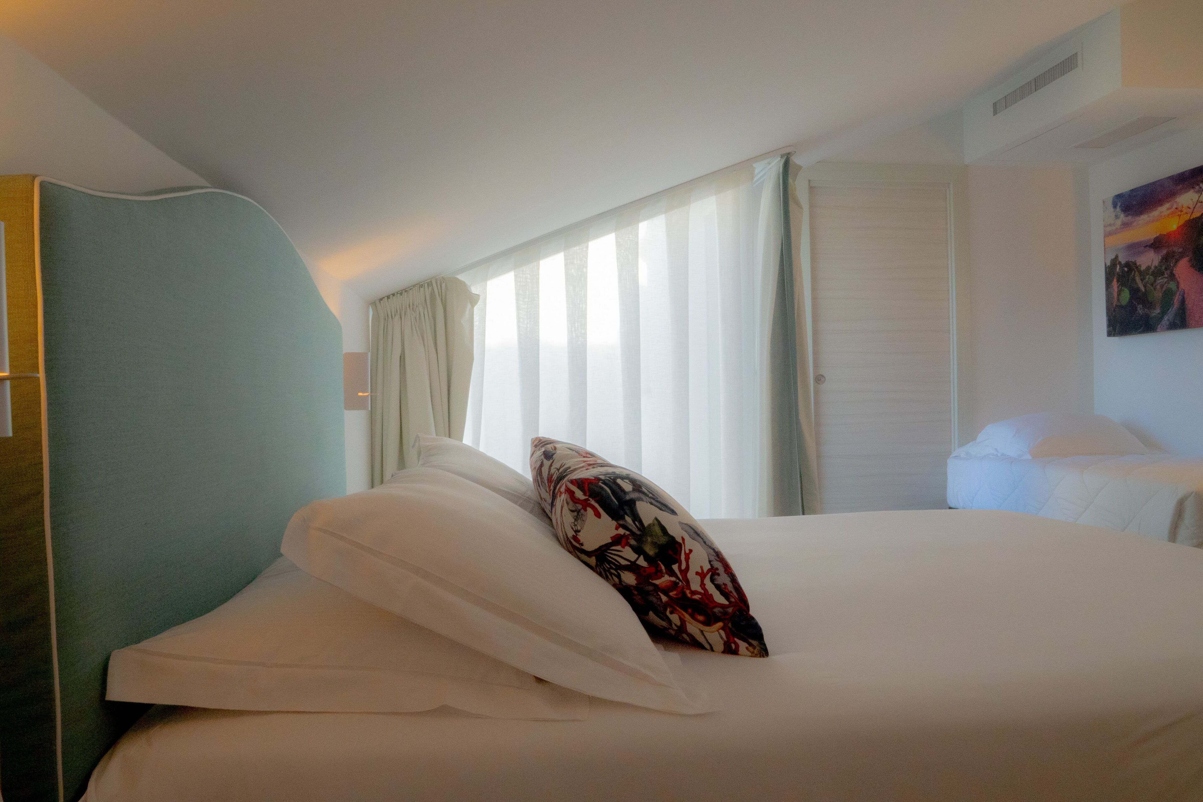 A hotel room with a bed, a headboard, a window with curtains, and a painting on the wall.