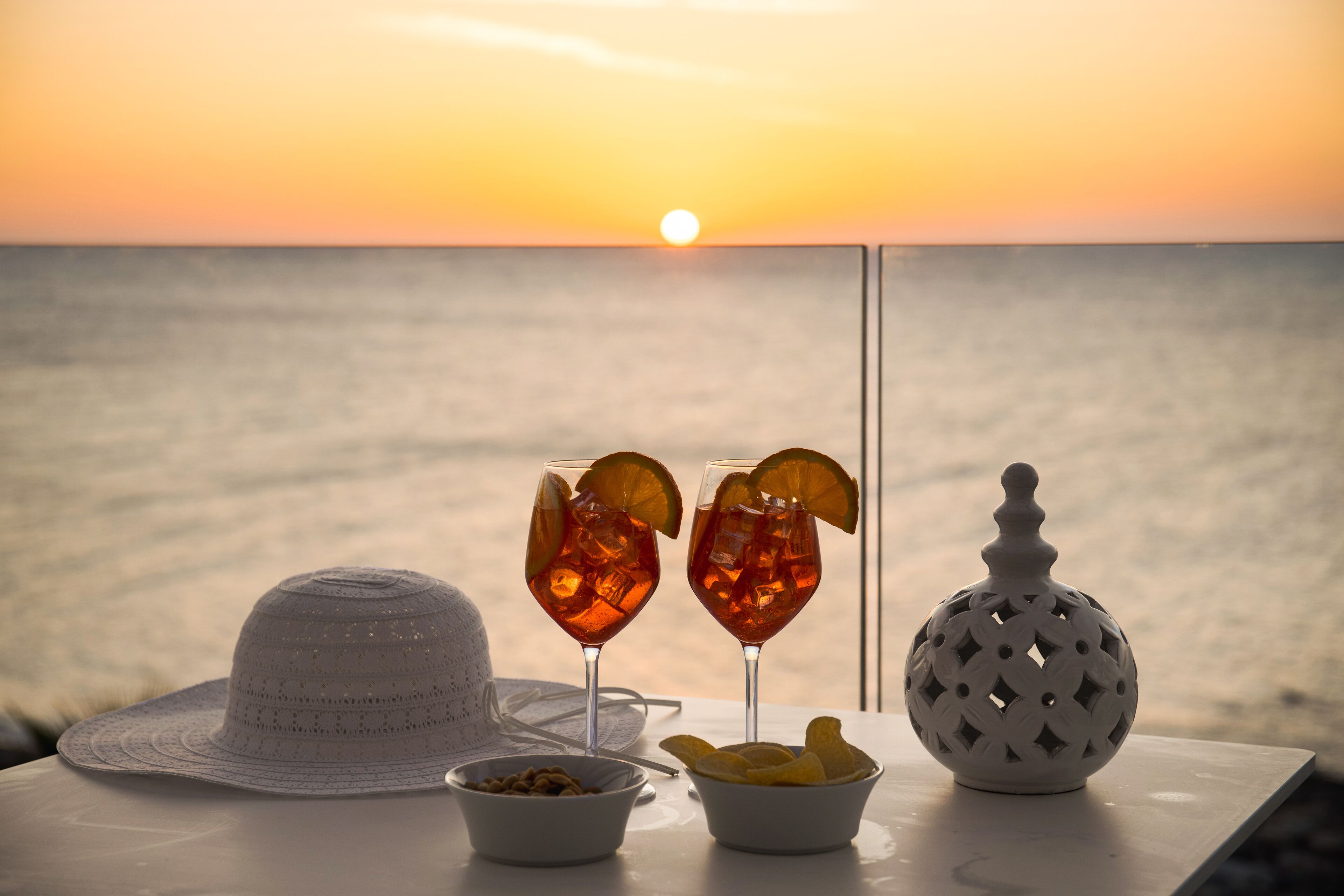 Una mesa con dos vasos de una bebida, un sombrero, un cuenco de bocadillos y un adorno, frente a una vista del atardecer.