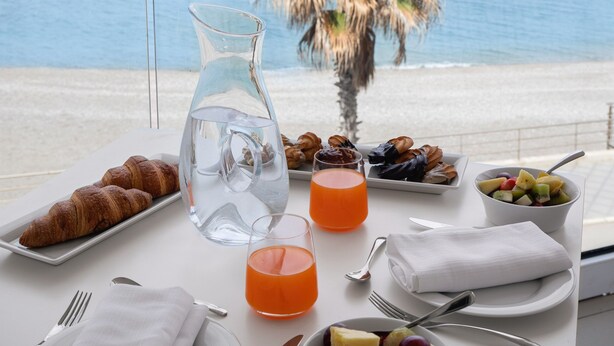 A breakfast table with croissants, fruit, and orange juice, overlooking a beach with palm trees and the sea. - Seaside Hotel (Capo d'Orlando)