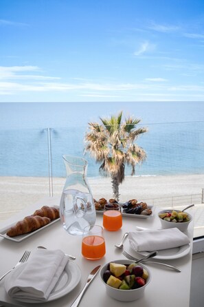 Una mesa de desayuno con croissants, fruta y jugo de naranja, con vista a una playa con palmeras y el mar.
