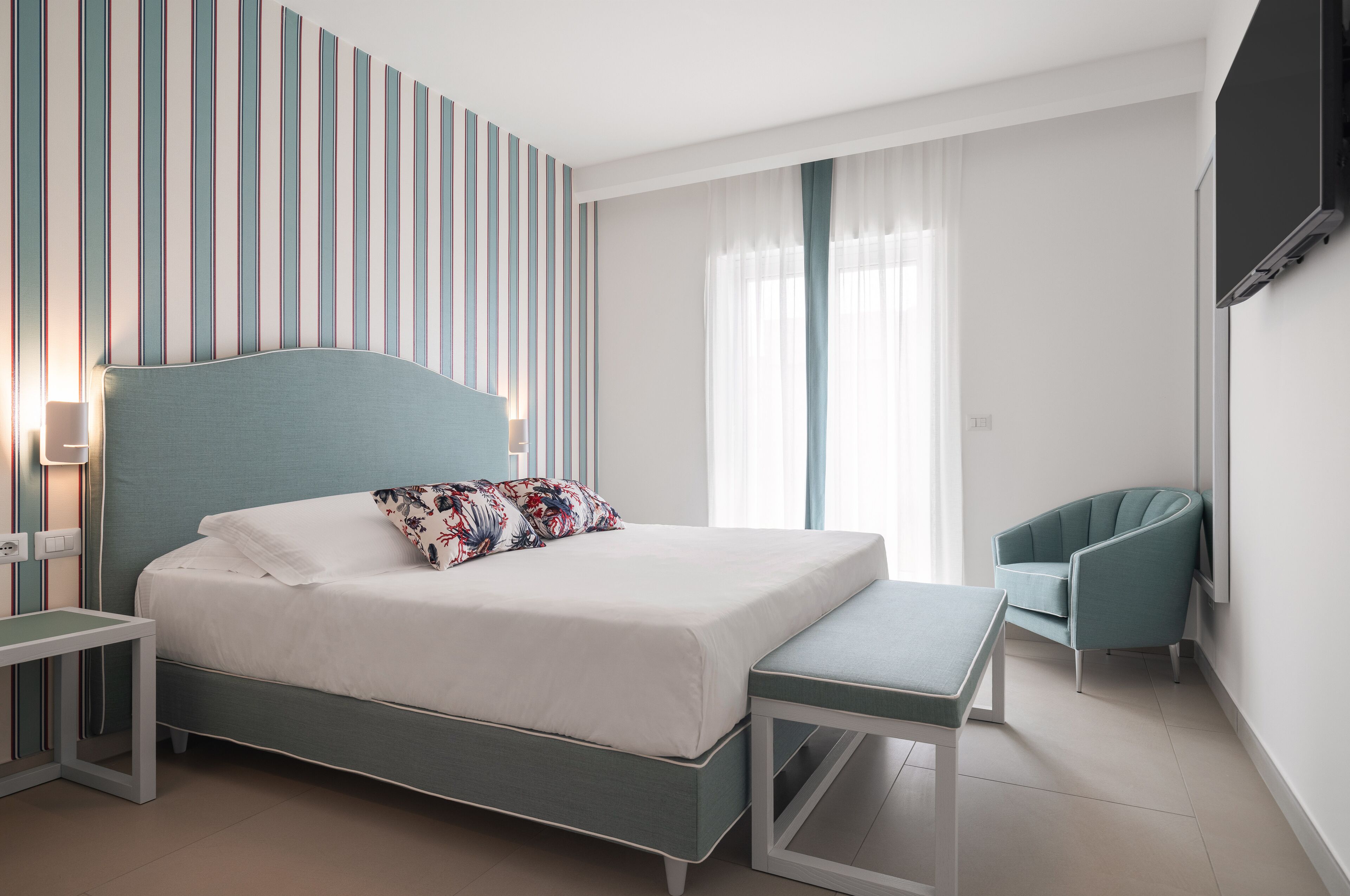 A modern bedroom with a bed, bench, chair, and a TV mounted on the wall.