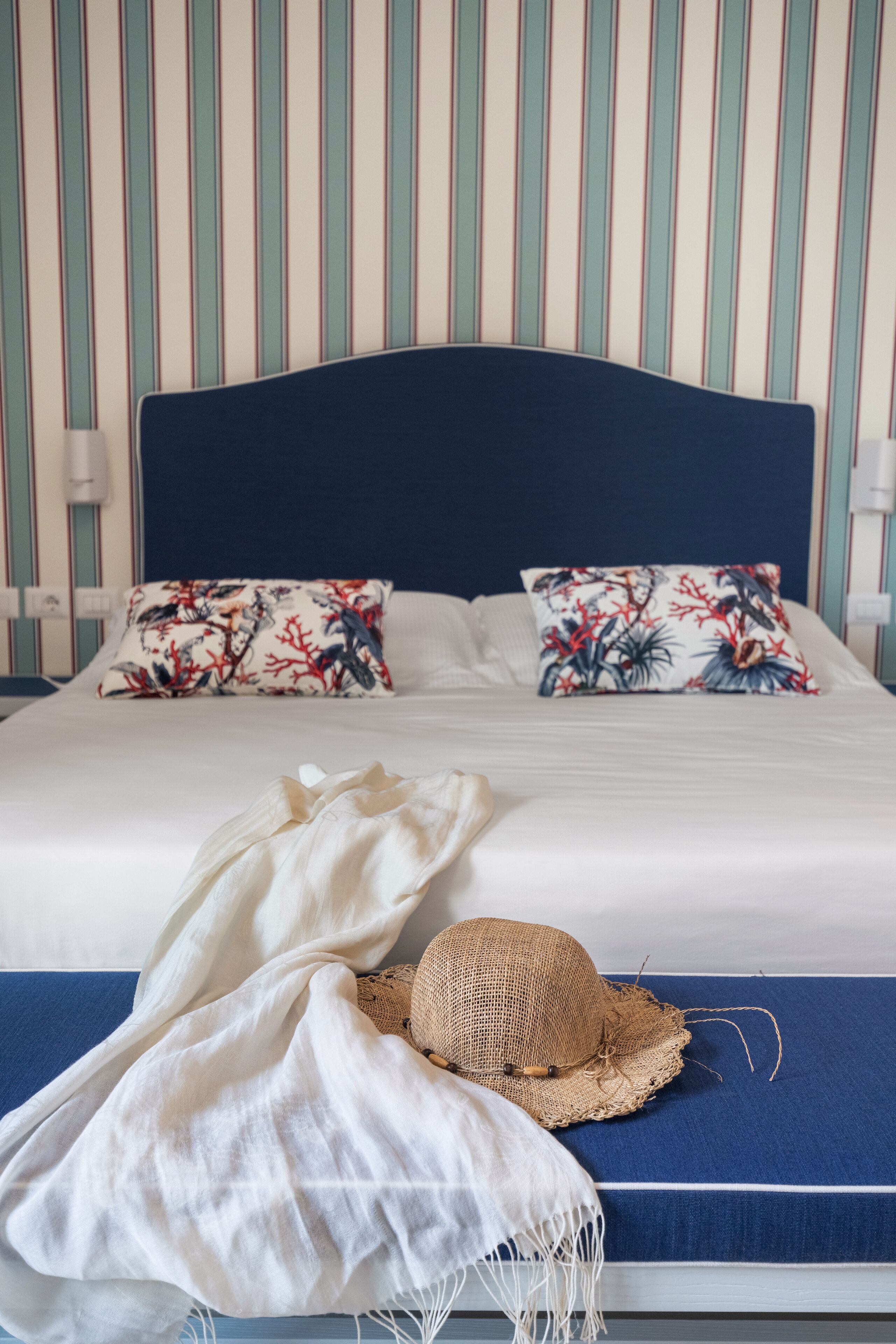 A bed with a blue headboard, two floral pillows, a straw hat, and a white throw blanket.