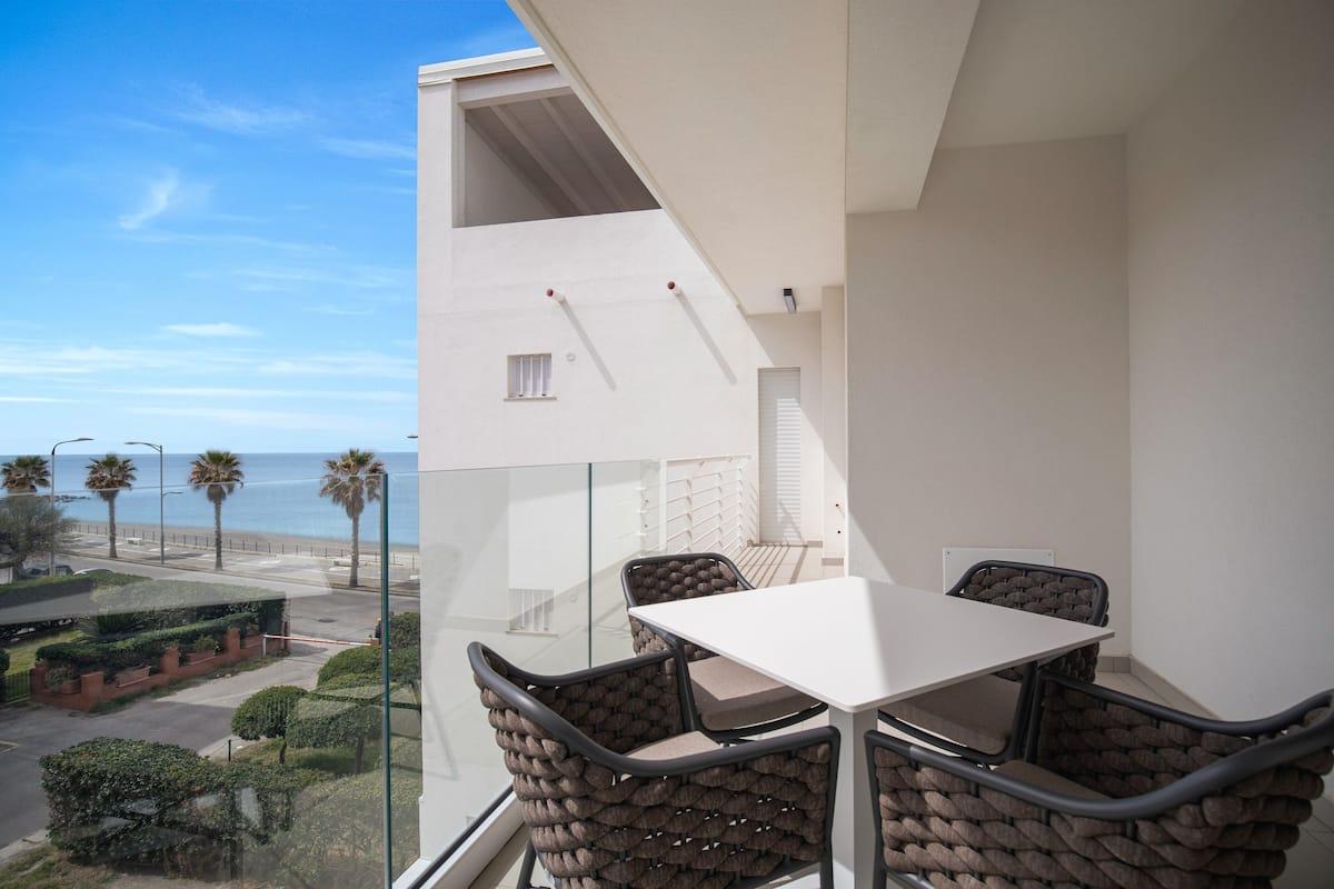 A balcony with a white table and chairs, overlooking a coastal street with palm trees and a clear sky.