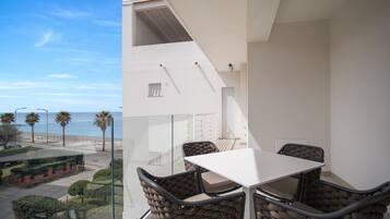 A balcony with a white table and chairs, overlooking a coastal street with palm trees and a clear sky.