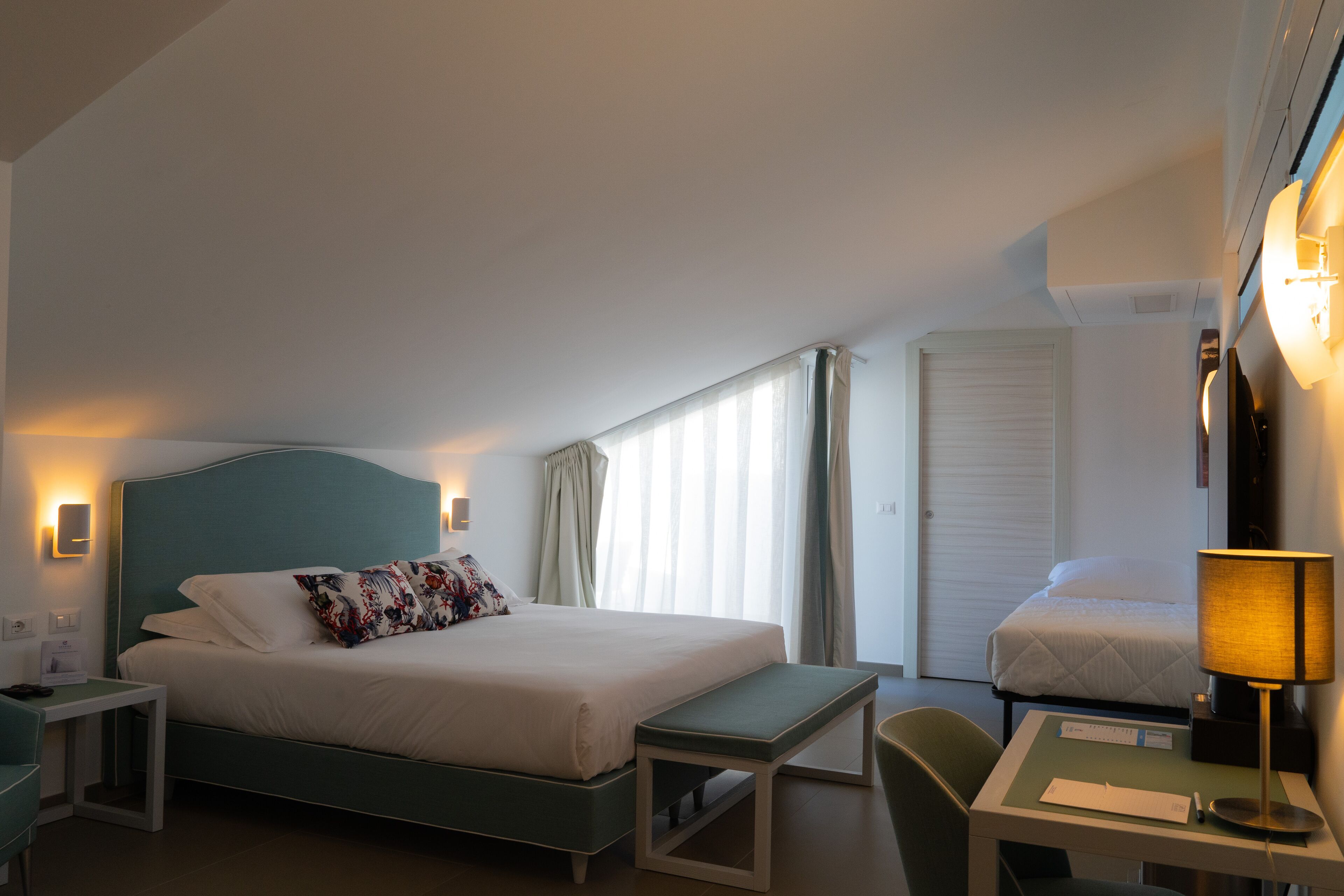 A hotel room with a large bed, a smaller bed, a desk with a lamp, a bench, and a window with curtains.