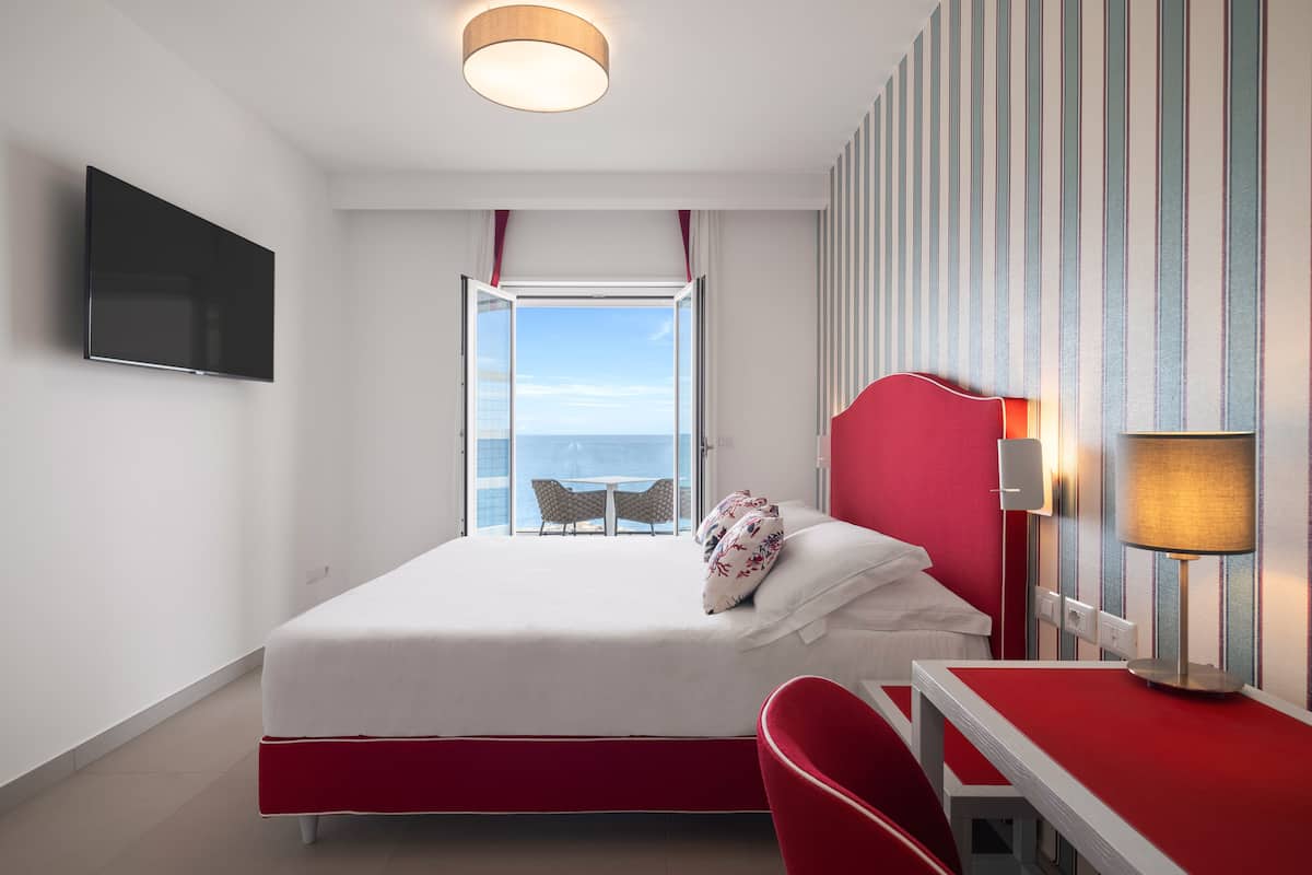 A hotel room with a bed, a desk, a chair, a TV, and a window with a view of the sea.
