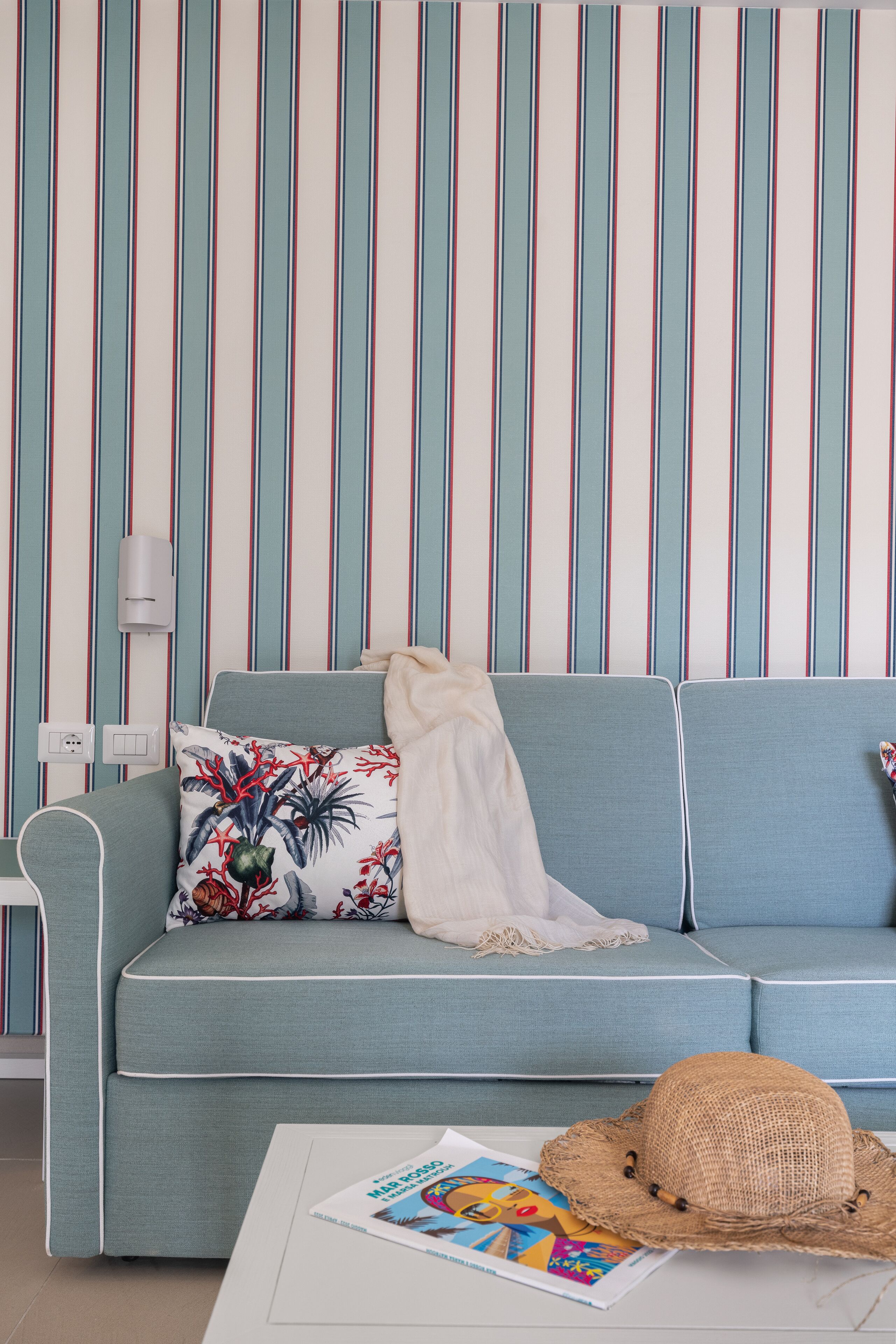 Een kamer met een lichtblauwe bank, een kussen met een patroon, een witte plaid, een strooien hoed en een tijdschrift op een witte tafel.