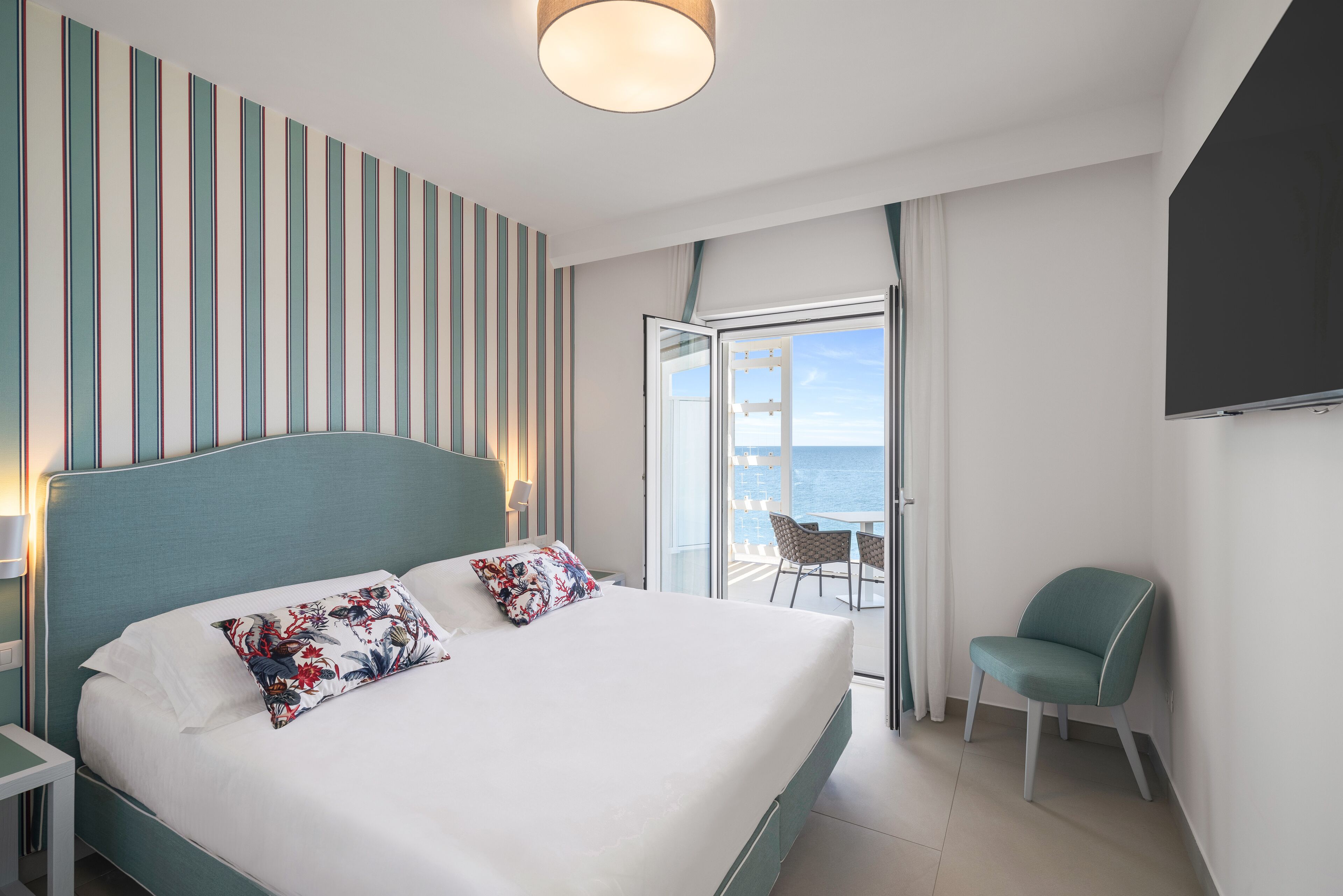 A modern hotel room with a large bed, a view of the sea, a balcony with a table and chairs, and a TV mounted on the wall.