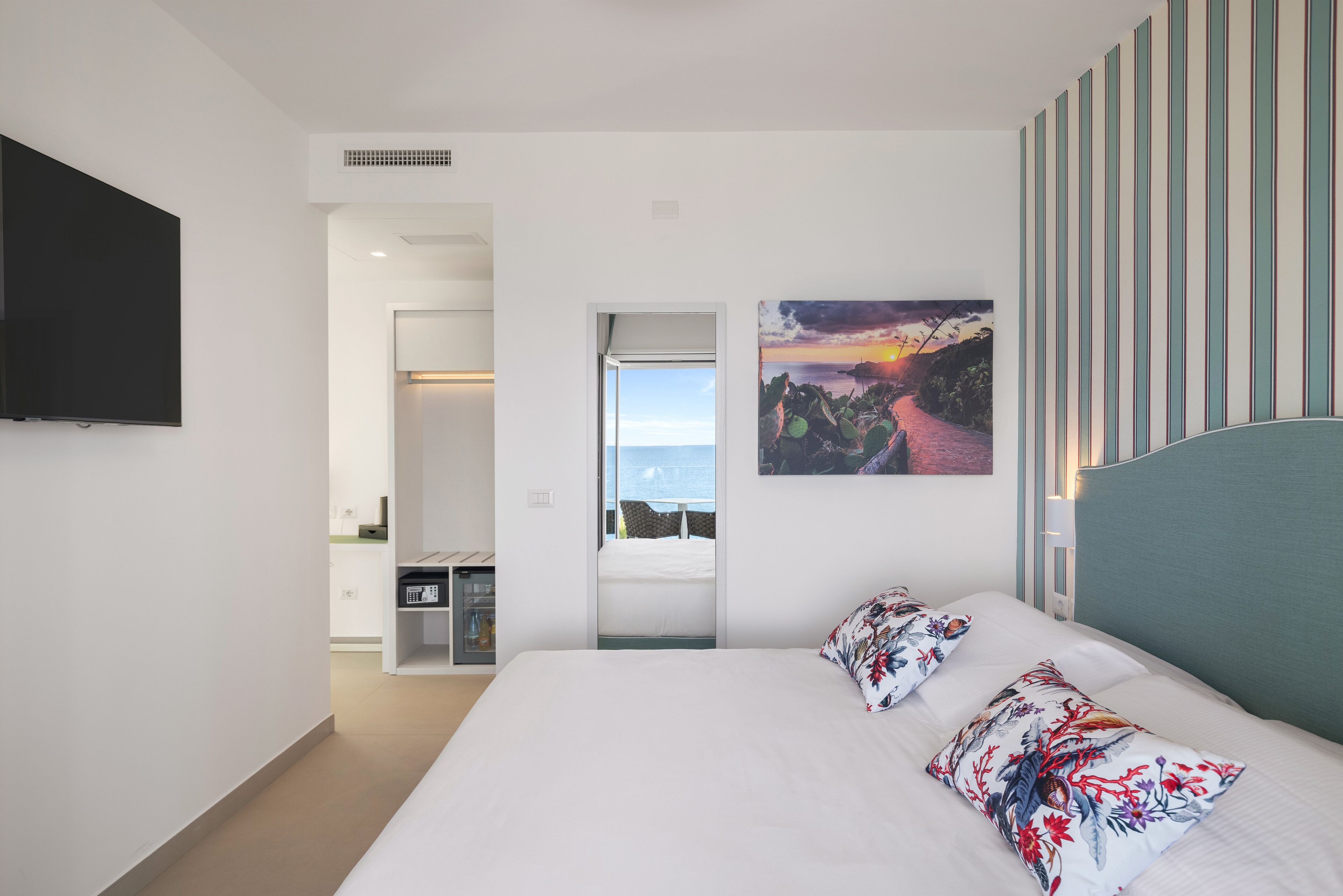 A modern hotel room with a large bed, a flat-screen TV mounted on the wall, a painting of a sunset, and a view through an open door.