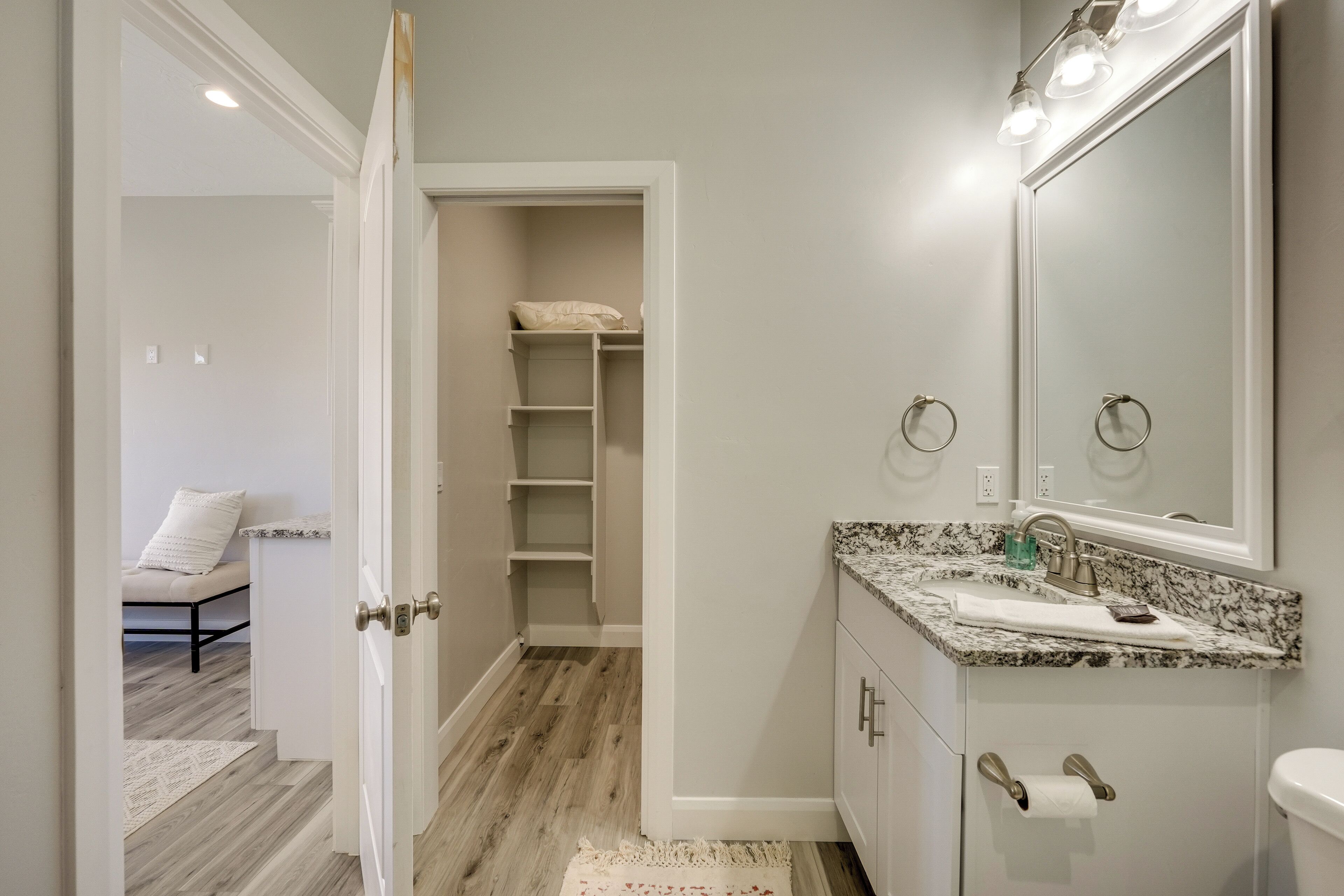 A bathroom with a vanity, sink, and mirror. There is a walk-in closet with shelves and a bed in the adjacent room.