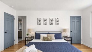 A bedroom with a large bed, two bedside tables, a nightstand, a door, and a wall with four framed artworks.