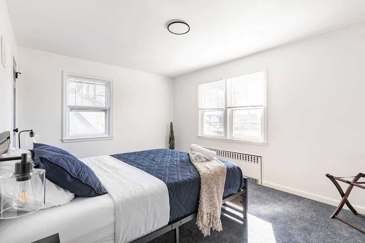 Un dormitorio con una cama, un radiador, un escritorio con una lámpara y dos ventanas.