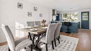 A dining area with a wooden table, grey upholstered chairs, and a vase of flowers. There are framed botanical prints on the wall.
