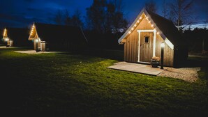 Eine kleine Holzhütte mit Veranda und Lichterkette, umgeben von Rasen und Bäumen.