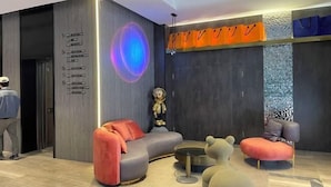 Eine moderne Lobby mit einem gebogenen grauen Sofa, einem runden Couchtisch und einem ottmanischen Hocker in Bärenform. Auf einem Sockel steht eine Statue, und eine Person ist im Hintergrund zu sehen.