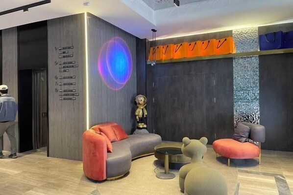A modern lobby with a curved grey sofa, a round coffee table, and a bear-shaped ottoman. There is a statue on a pedestal, and a person is visible in the background.