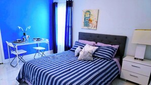Un dormitorio con colcha a rayas, cabecera, mesita de noche, lámpara y un cuadro en la pared.