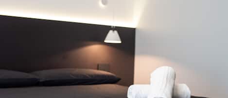 A neatly made bed with a dark headboard, a single white towel, and a pendant light above.