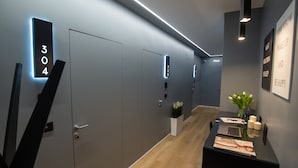 A modern hallway with numbered doors, a wooden floor, a desk with a laptop, and a vase of flowers.