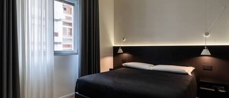 Un dormitorio moderno con una cama grande, una mesita de noche, una lámpara de pared y una ventana con cortinas.